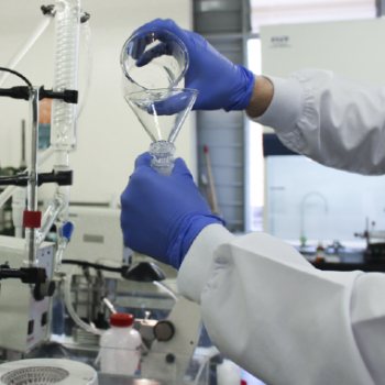 En la imagen se puede observar dos manos con guantes azules, ideales para el trabajo en laboratorio, están vertiendo de un frasco a otro una sustancia trabajada o elaborada en el laboratorio. Para la Escuela de Química es importante dar a conocer sus grupos de investigación.